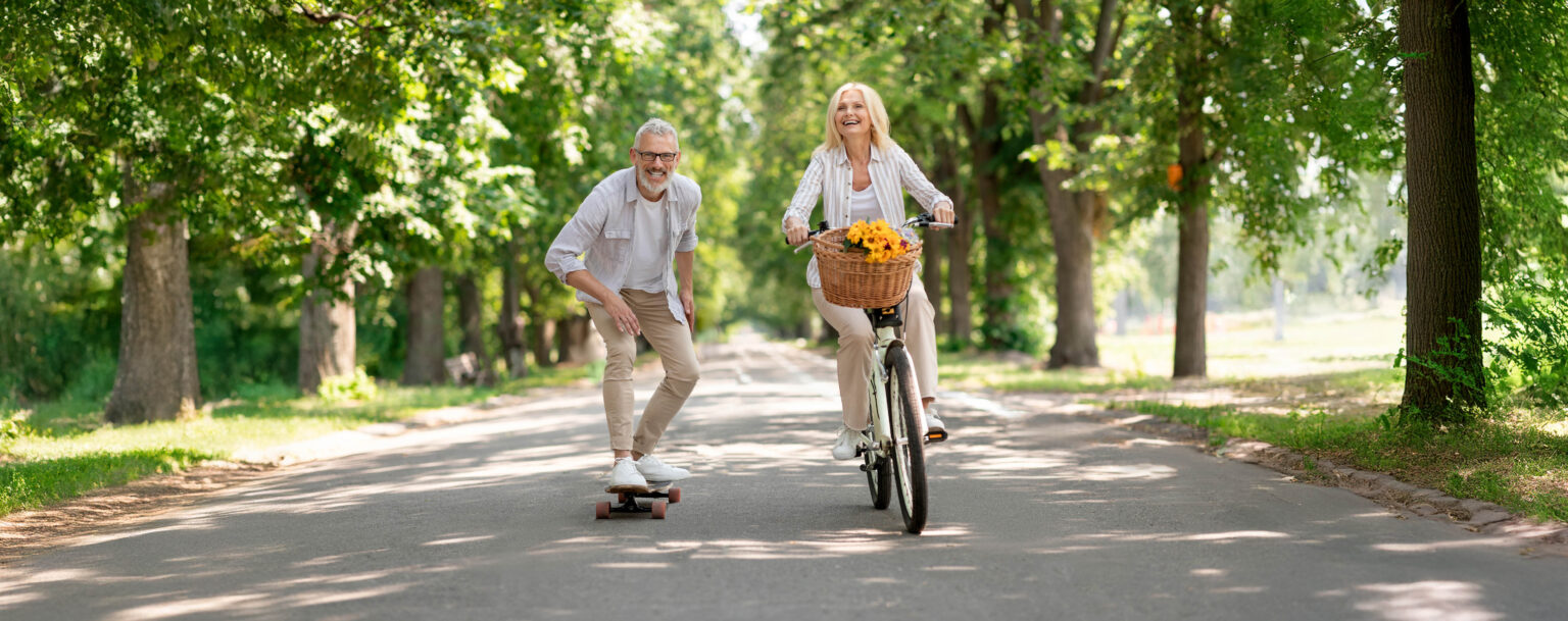 Fröhliche Rentner fahren eine Allee entlang. Der Mann fährt auf einem Skateboard und die Frau auf einem Fahrrad
